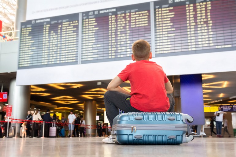 Lonely child sitting on a suitcase and waiting for departure at the airport looking at the arrivals board.