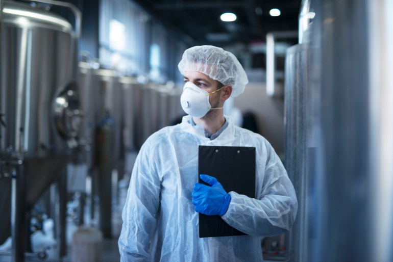 Technologist in protective white suit with hairnet and mask standing in food factory.