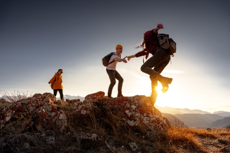 Group of young hikers with backpacks walks with backpacks and helps each other in climbing in sunset mountains