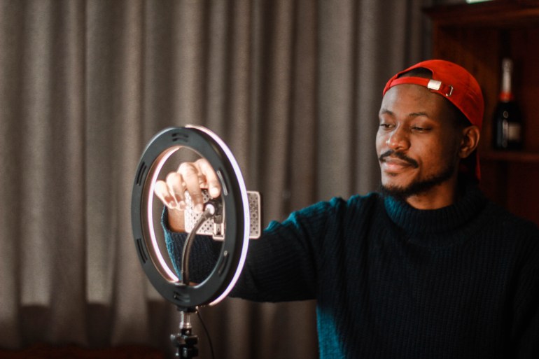 Influencer from South Africa vlogging while self-isolating. He is wearing a red cap. He is using a ring-light and a smartphone.