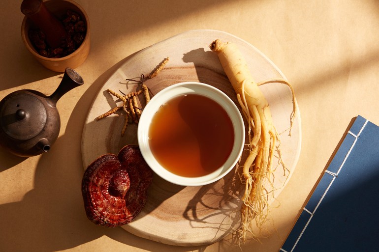 A bowl of tonic made from ginseng, Ganoderma lucidum, and cordyceps is displayed on a wooden platform. Books, teapots, and mortars of dried herbs on the golden paper background; Shutterstock ID 2218536173; purchase_order: aj; job: ; client: ; other: