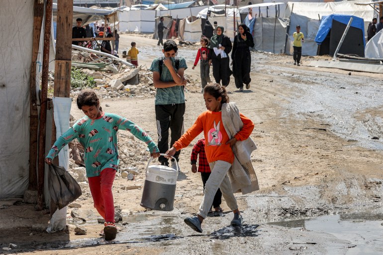 Two girls carry together a water cooler as they walk past tent shelters in the Nasr neighbourhood in western Gaza City on April 15, 2025.