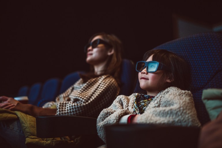 Mom & daughter watching movie with 3D glasses in cinema hall at movie theatre.