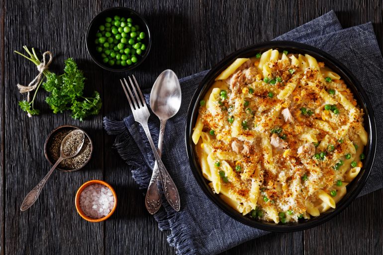 تونة Tuna Mornay, Creamy Tuna Casserole Penne Pasta Bake in baking dish on dark wooden table with fork and spoon, horizontal view from above, flat lay
