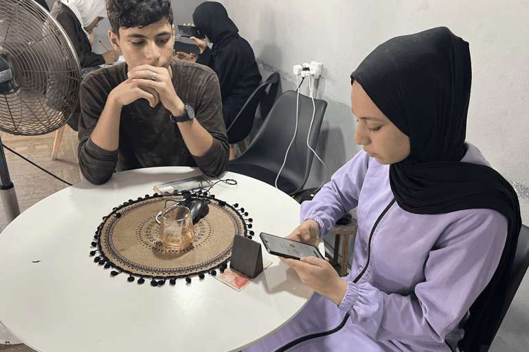 الطالبة ليلى البطش تحدق في شاشة هاتفها بخيبة أمل بعد إعلان تأجيل الاختبار.